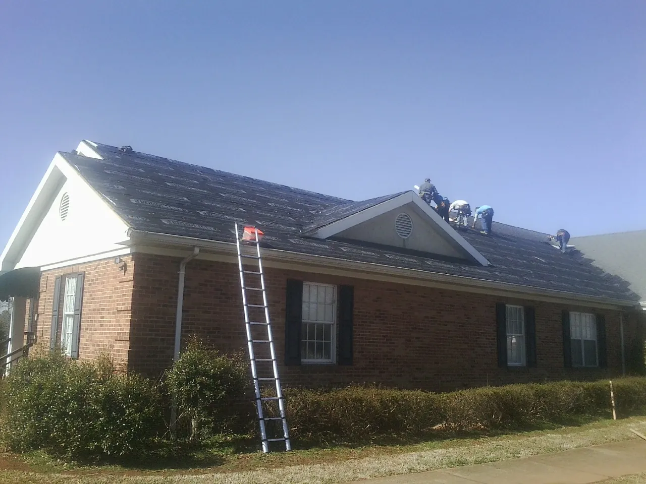 Crew tearing shingles off a brick ranch roof with a ladder set — Metal Roof Repair work by our crew in Colliers