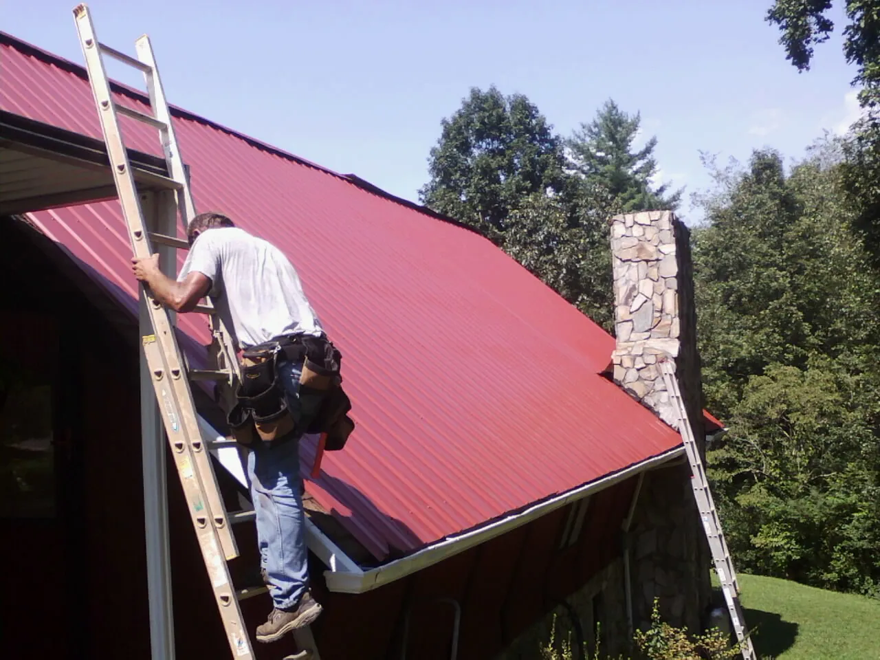 Roofer on a ladder beside a red metal roof and stone chimney — Soffit & Fascia Repair by Roofing Brothers in Colliers